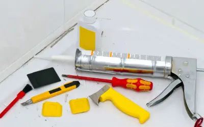 Werkzeuge zum Silikon von Fliesen entfernen: Kartuschenpresse, Cutter, Schaber, Bürste und eine Flasche mit Reiniger.