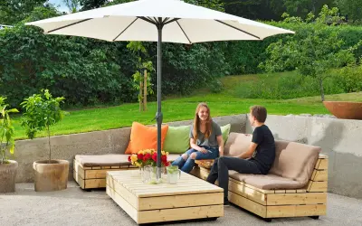 Två personer njuter av solen under ett parasoll vid en hemmabyggd loungesoffa i trä.