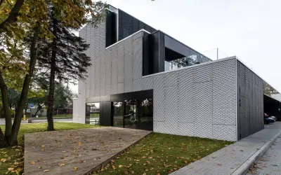 Nowoczesny budynek z metalicznym ryzalitem, przeszklonym wejściem i drewnianym tarasem porośniętym jesiennymi liśćmi.