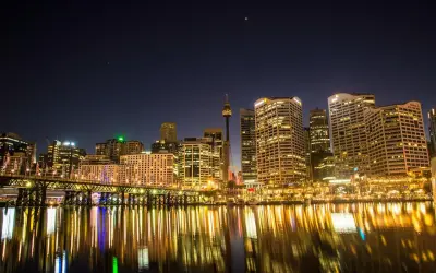 Sydney nocą, z mostem i odbijającymi się w wodzie światłami. Zastanawiasz się, ile się leci do Australii, patrząc na ten widok?