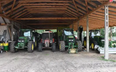 Kilka zielonych i jeden czerwony traktor stoją pod drewnianą wiatą. To pewnie ich tymczasowe schronienie, bo taka wiata bez pozwolenia to nie najlepszy pomysł.