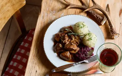 Piatto di cinghiale in dolceforte con canederli e cavolo rosso. Un bicchiere di vino rosso e un palco di cervo completano la scena rustica.