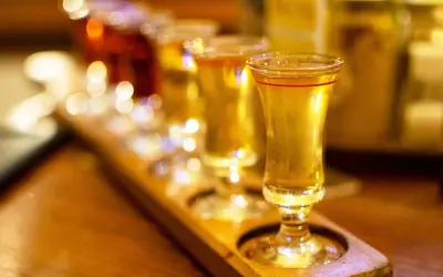 Degustacja drinków z syropem z czarnego bzu. Złociste płyny w kieliszkach na drewnianej desce, w tle rozmyte światła.