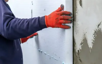 Ręce w pomarańczowych rękawicach przytwierdzają płyty styropianowe do ściany, wykonując ocieplenie stropu styropianem.