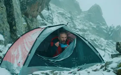 Mężczyzna w namiocie, jedzący posiłek, w zimowej scenerii górskiej. Scena jak z filmów wspinaczkowych.