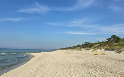 Słoneczny dzień nad zalewem kamieńskim. Szeroka, piaszczysta plaża z łagodnymi falami i zielonymi wydmami w tle. Kilka osób odpoczywa na piasku.