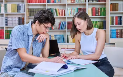 Dos personas jóvenes estudian juntas en una biblioteca, rodeadas de estanterías llenas de libros.