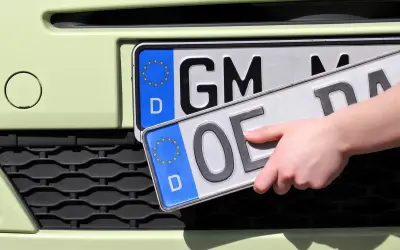 Dłoń trzyma tablice rejestracyjne państw. Jedna z nich ma oznaczenie "GM", druga "OE".