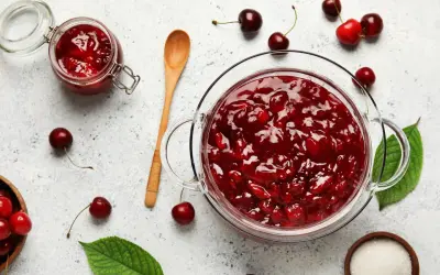 Un bocal et un bol en verre débordent de confiture de cerises maison. Des cerises fraîches et des feuilles vertes parsèment la surface.