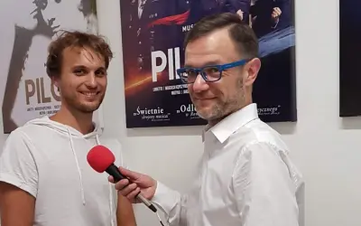 Mężczyzna w białej koszuli z niebieskimi okularami trzyma mikrofon, udziela wywiadu młodemu, uśmiechniętemu czarneckiemu aktorowi na tle plakatów teatralnych.