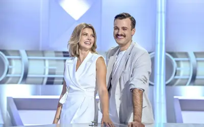 Uśmiechnięci prowadzący koło fortuny, kobieta w białej sukience i mężczyzna w jasnej marynarce, stoją przy stole w studiu.