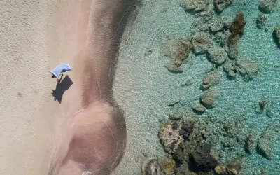 En person med en blå handduk på en rosa strand på Kreta. Klart turkost vatten med stenar.