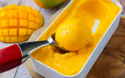 Une cuillère prélève une boule de sorbet mangue crémeux d'une boîte. Des morceaux de mangue fraîche sont visibles à côté.