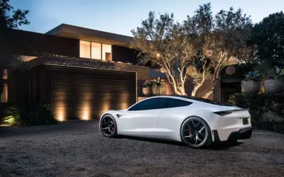 Biały roadster Tesla stoi na żwirowym podjeździe przed nowoczesnym domem.