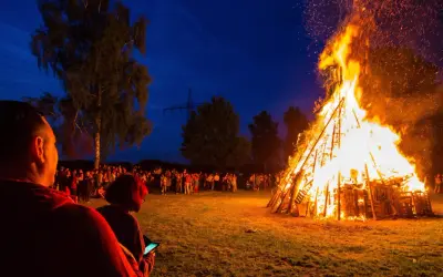 Menschen versammeln sich, um die Sommersonnenwende zu feiern. Ein riesiger Holzstapel brennt lichterloh im Abendhimmel, Funken steigen auf.