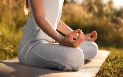 En person sitter i lotusställning på en matta utomhus, med händerna i meditation. Solen lyser genom gräset och skapar en varm atmosfär för morgonmeditation.
