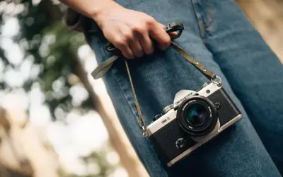 Dłoń trzyma aparat OM-3, przypominając o czasach, kiedy powstał aparat fotograficzny.