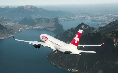 Samolot linii Helvetic Airways z charakterystycznym czerwonym ogonem i białym krzyżem leci nad malowniczym jeziorem i górami.