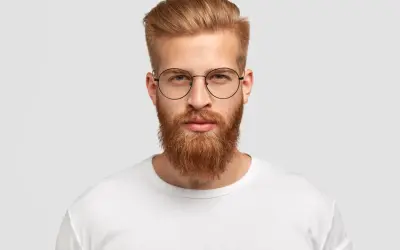 Un homme au visage rectangulaire, barbe rousse et lunettes rondes, porte un t-shirt blanc.