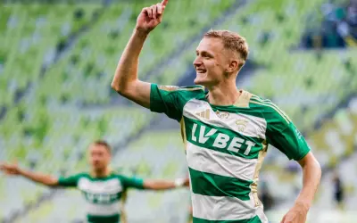 Radosny środkowy napastnik w zielono-białej koszulce świętuje bramkę na tle zielonych trybun stadionu.