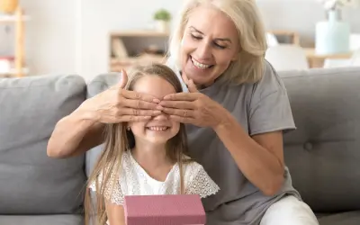 Babcia zasłania oczy wnuczce, która trzyma prezent. Radosna niespodzianka z okazji imienin Nikodema.