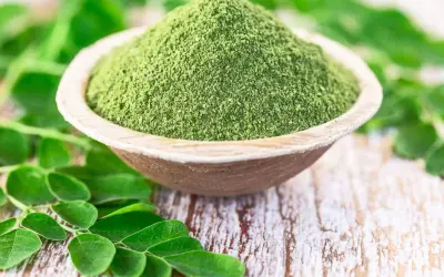 Moringa w proszku w drewnianej misce, otoczona świeżymi liśćmi. Zanim zaczniesz stosować, sprawdź moringa przeciwwskazania.