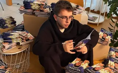 Młody mężczyzna w okularach siedzi otoczony stosami czasopism z autorskimi krzyżówkami. Zarabianie na układaniu krzyżówek do czasopism to jego sposób na życie.