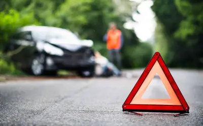 Triangle de signalisation sur la route, près d'une voiture accidentée où une personne gît au sol. Un homme en gilet orange se tient debout. Scène évoquant un homicide involontaire.