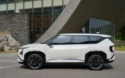 Nowy, biały suv Kia EV9 stoi zaparkowany przed nowoczesnym budynkiem z kamienną fasadą i dużymi oknami.