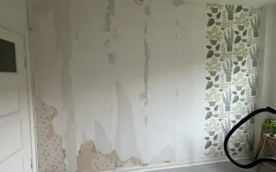 Väggen är delvis avskalad, en del tapet sitter kvar. En ny tapet med gröna växter är på väg att måla på tapet eller riva.