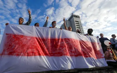 Ludzie trzymają biało-czerwoną flagę, symbol wolności Białorusi. Niebo z chmurami nad budynkiem.