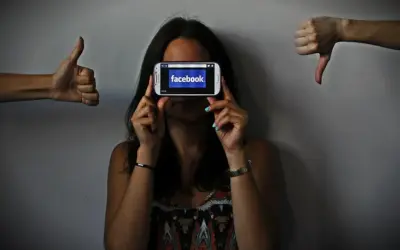 Una persona oculta su rostro tras un móvil con Facebook. A los lados, pulgares arriba y abajo, reflejando el impacto de las redes sociales y la autoestima.