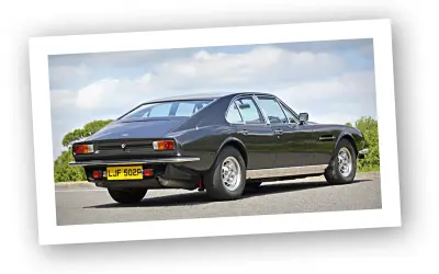 Klasyczny Aston Martin Lagonda w ciemnym kolorze, widziany z tyłu, na tle błękitnego nieba z chmurami.