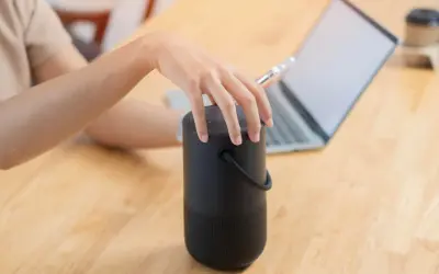 Dłoń dotyka czarnego głośnika, obok laptopa. Dowiedz się, jak podłączyć głośniki do komputera, aby cieszyć się lepszym dźwiękiem.