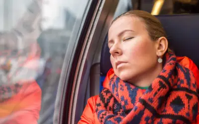 Kobieta w pomarańczowej kurtce i wzorzystym szaliku odpoczywa w pociągu, wyraźnie odczuwając zmęczenie.