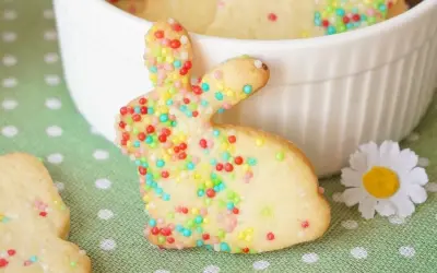 Bunte Osterkekse zum Ausstechen, verziert mit bunten Streuseln, liegen auf einem grünen Tupfentuch. Ein süßer Osterhase steht im Vordergrund.