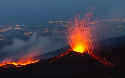Spektakularna erupcja Etny nocą. Wulkan wyrzuca strumienie lawy i iskier na tle rozświetlonego miasta.