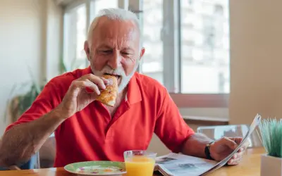 Un homme âgé mange un croissant en lisant le journal, un verre de jus d'orange à côté. Une scène qui illustre les besoins nutritionnels de la personne âgée.