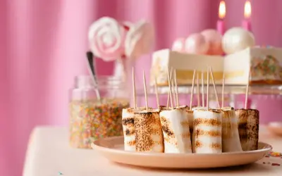Grillade marshmallows på spett, perfekt fika till barnkalas. En tårta och strössel i bakgrunden.