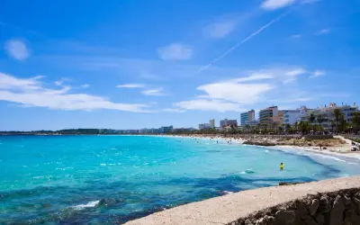En livlig Cala Millor Beach med turkost vatten, solbadande människor och hotell längs kusten under en klarblå himmel.