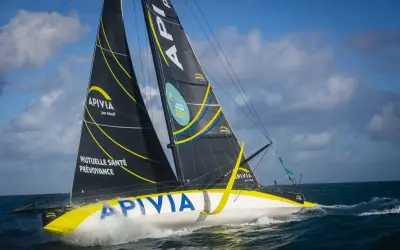 Le voilier Apivia, skippé par Alex Thomson, fend les vagues sous un ciel bleu et nuageux.