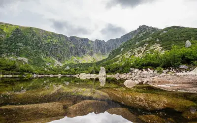 Spokojne, górskie jezioro odbija skaliste zbocza i chmury. W oddali widać skalne formacje, a na brzegu leżą kamienie. To piękne, górskie krajobrazy, które przypominają o potędze natury i jej śnieżnych stawkach.