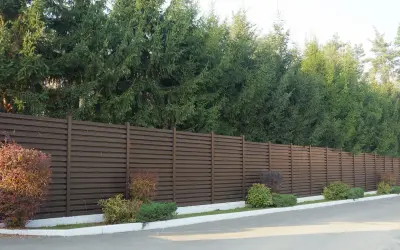 Ciemnobrązowy płot lamelowy, za którym rosną wysokie świerki, stanowi świetne rozwiązanie, czym zasłonić płot od sąsiada.