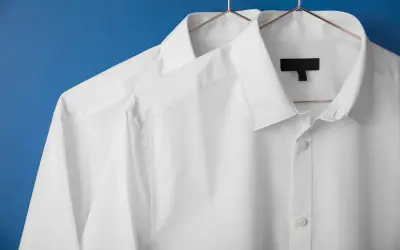 Två vita skjortor hänger på galgar mot en blå bakgrund. De ser ut som om de precis kommit från en **dry cleaner**, perfekt strukna och redo att bäras.