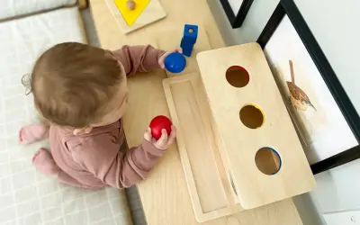 A baby explores Montessori activities for 10 month old baby, holding red and blue balls near a wooden drop box and shape puzzle.