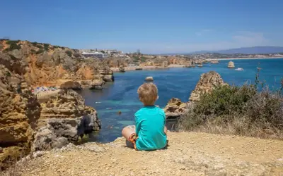Kleinkind blickt auf die Küste Portugals. Felsen, türkisfarbenes Meer und eine Bucht mit Strand.