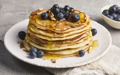 Stapel fluffiger High Protein Pancakes mit Blaubeeren und Mandelsplittern, beträufelt mit Honig.