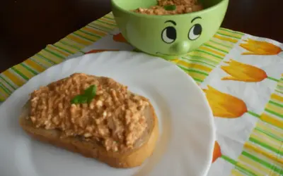 Pyszna pasta z makreli i twarogu na kromce chleba, ozdobiona listkiem bazylii. Obok miseczka z pastą i wesoła buźka.