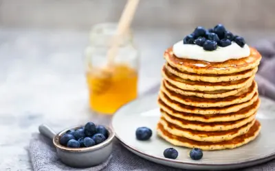 Pile de pancakes moelleux, garnis de crème et de myrtilles, avec un pot de miel en arrière-plan. Une recette pancake parfaite pour le petit-déjeuner.