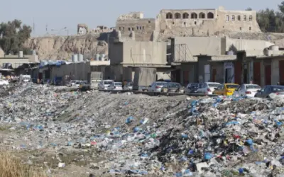 Müllberge säumen die Straßen von Kirkuk, Irak. Autos parken hinter den Geschäften, während im Hintergrund alte Gebäude auf einem Hügel stehen.